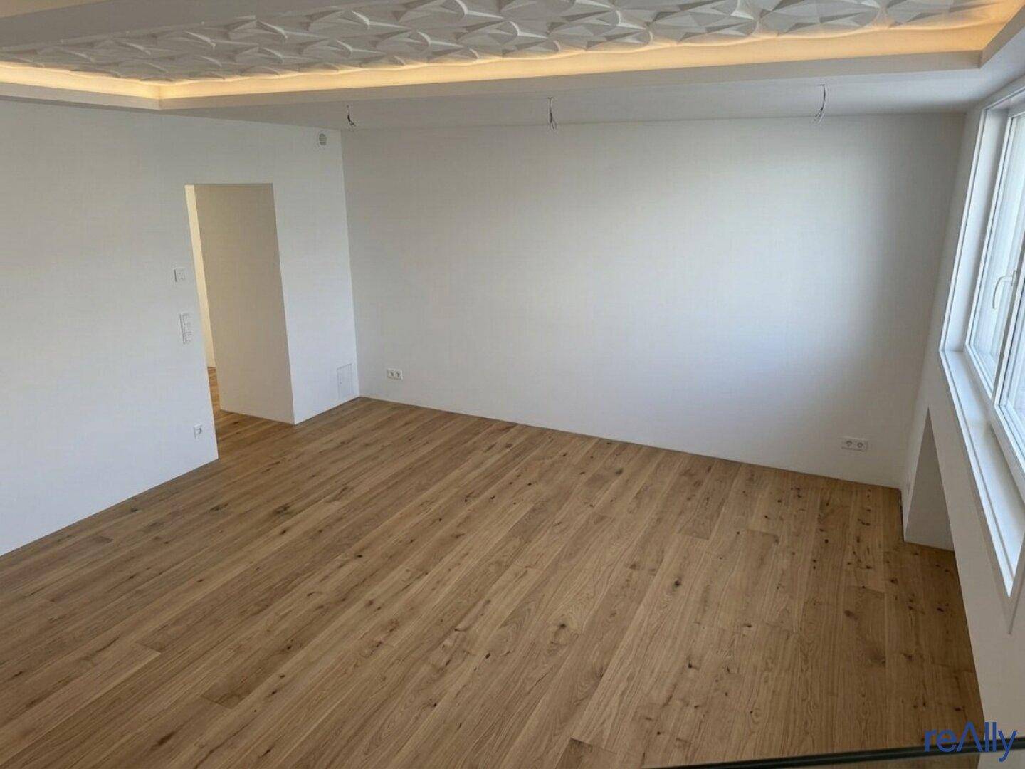 Heller, leerer Raum mit weißen Wänden, Holzboden und einer dekorativen Deckenverkleidung.