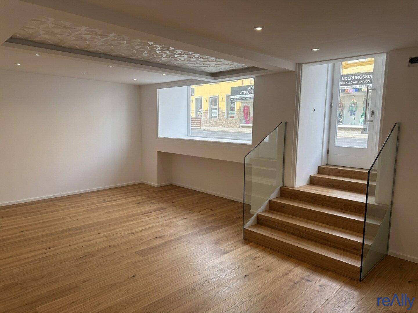 Heller, leerer Raum mit Holzboden, moderner Decke und einer Treppe mit Glasgeländer.