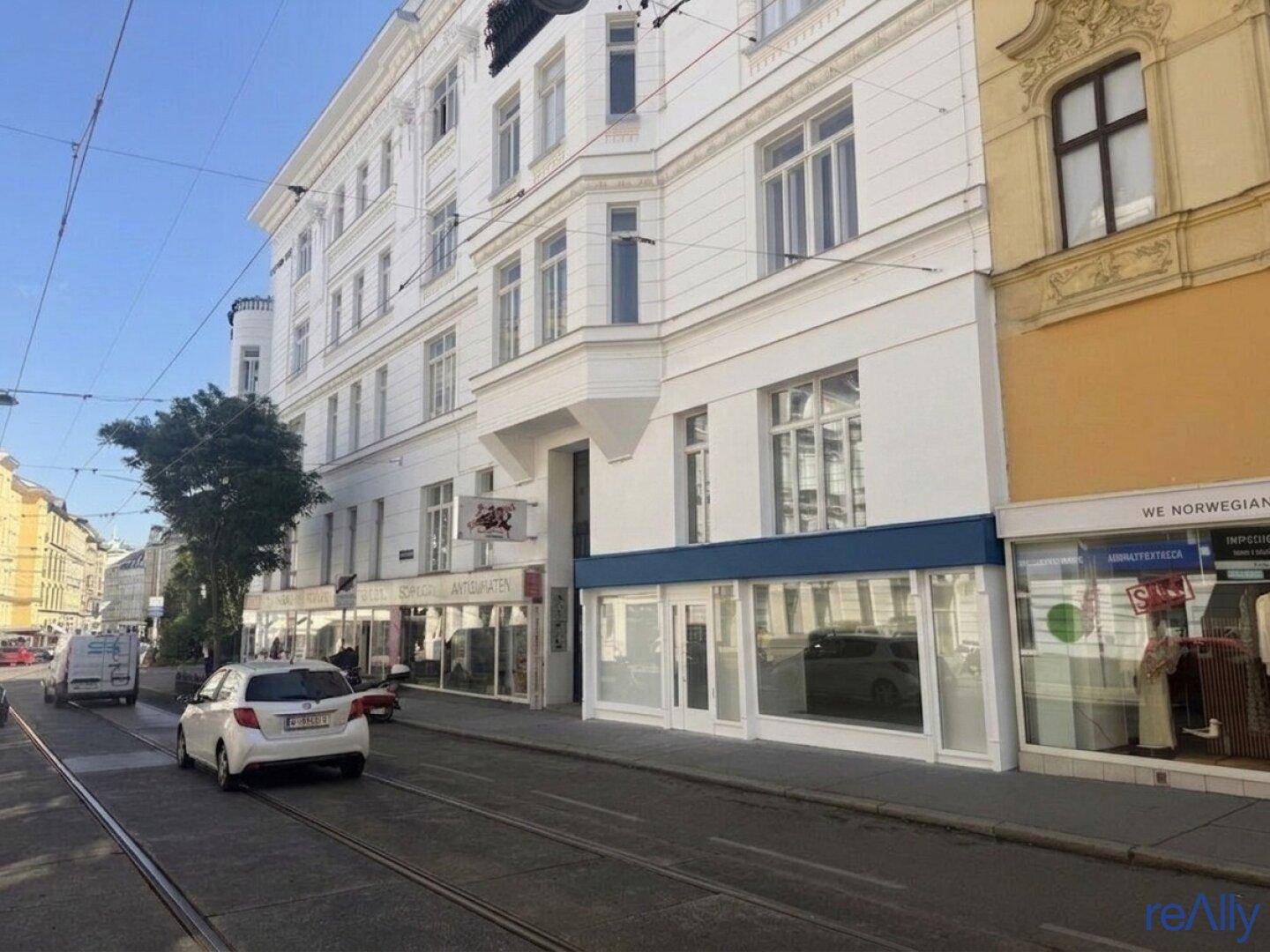 Gebäudeansicht an einer belebten Straße mit Geschäften im Erdgeschoss und Straßenbahnschienen.