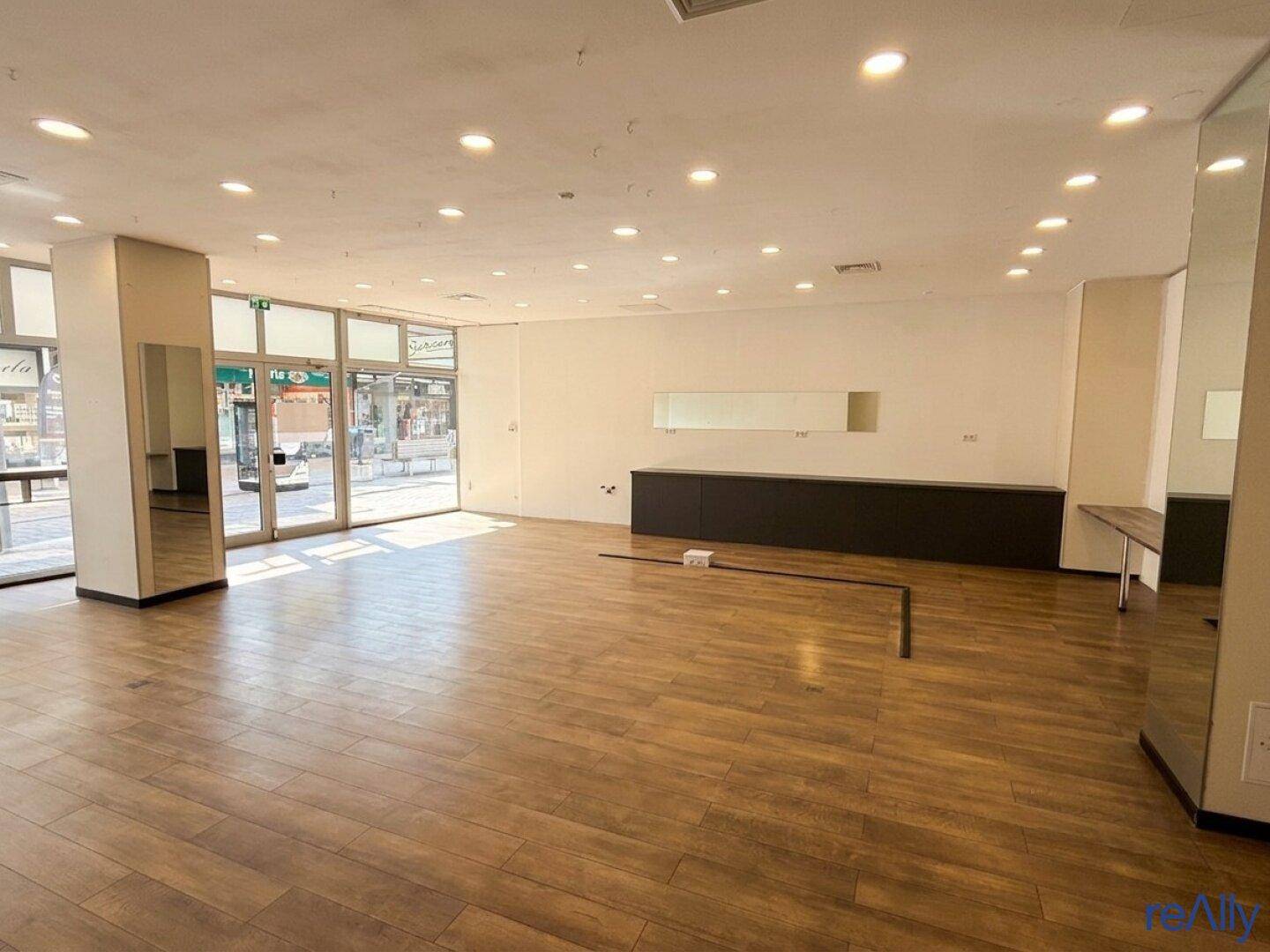 Offener Ladenbereich mit glänzendem Holzboden, moderner Deckenbeleuchtung und breiten Schaufenstern für optimale Präsentation.