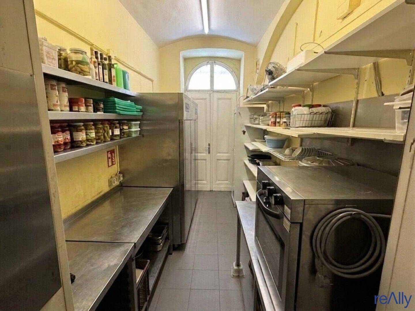Lagerraum der Gewerbeküche mit Regalen voller Vorräte, großem Kühlschrank und einem Fenster am Ende des Raumes.