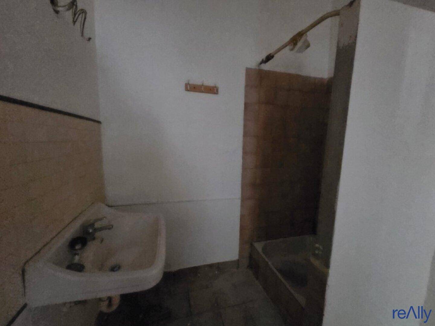 Einfaches Badezimmer mit Waschbecken und Duschbereich, stark renovierungsbedürftig.