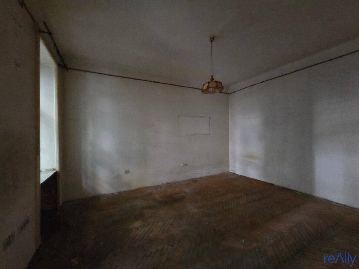 Geräumiges Zimmer mit abgenutztem Parkettboden und einfacher Deckenbeleuchtung, sanierungsbedürftig.