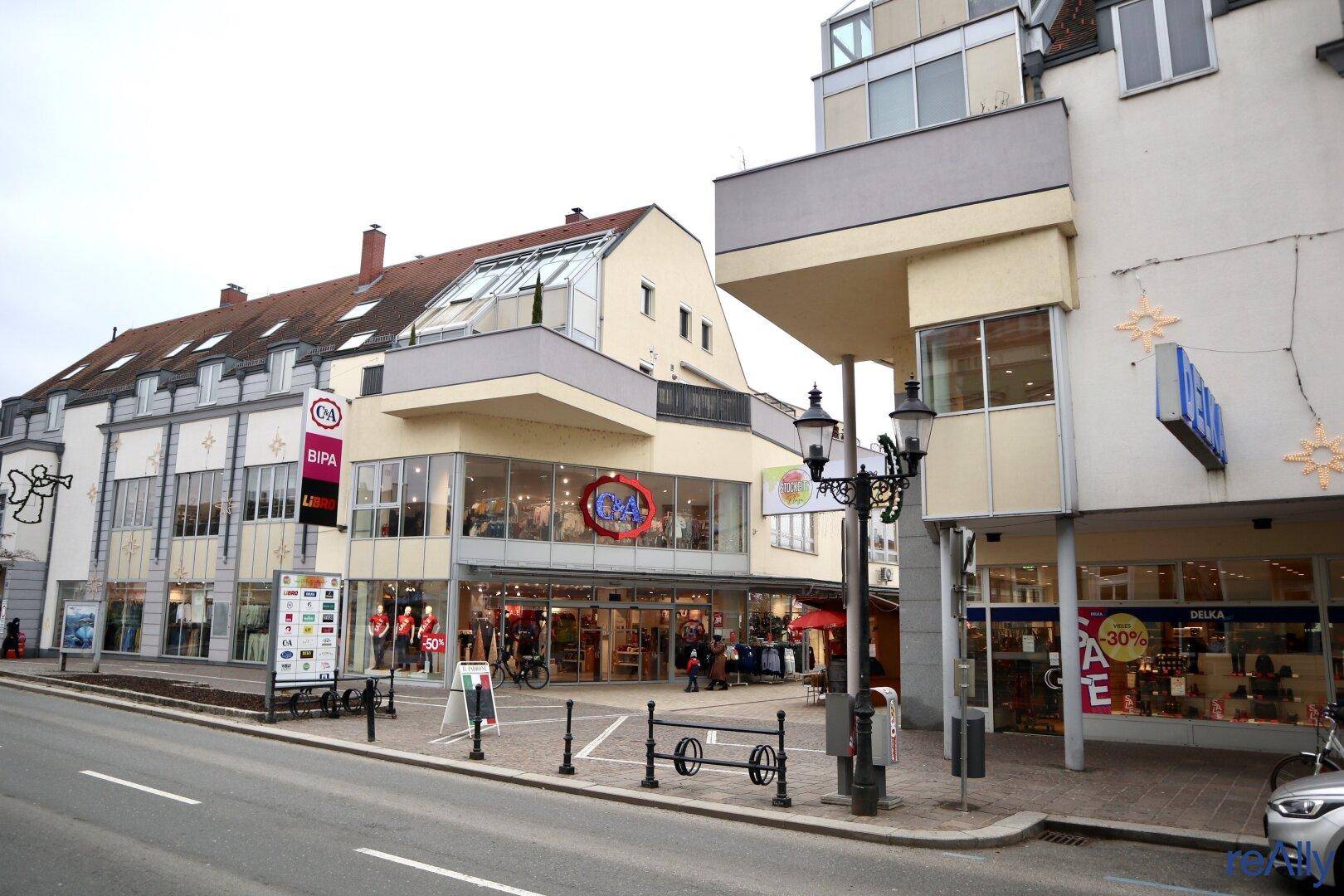 Straßenansicht mit mehreren Geschäften und Gebäuden, darunter C&A und BIPA, an einem bewölkten Tag.