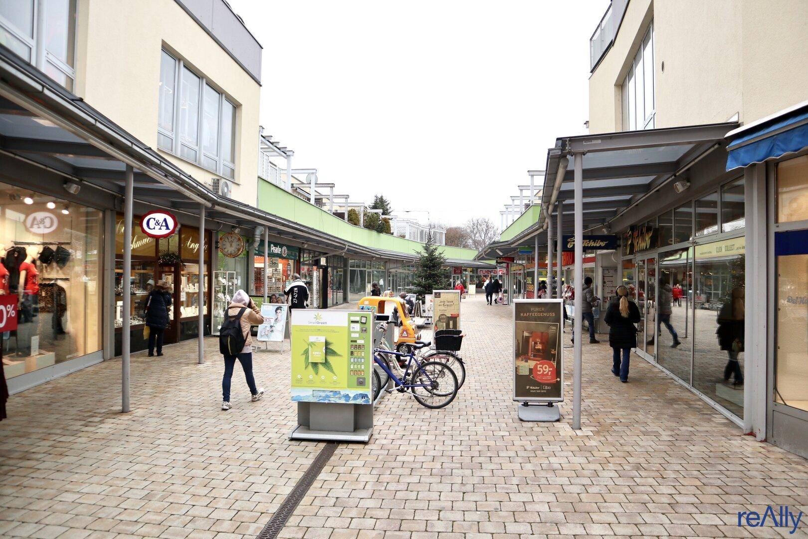Blick auf eine belebte Einkaufsstraße mit verschiedenen Geschäften und Passanten unter einem bewölkten Himmel.