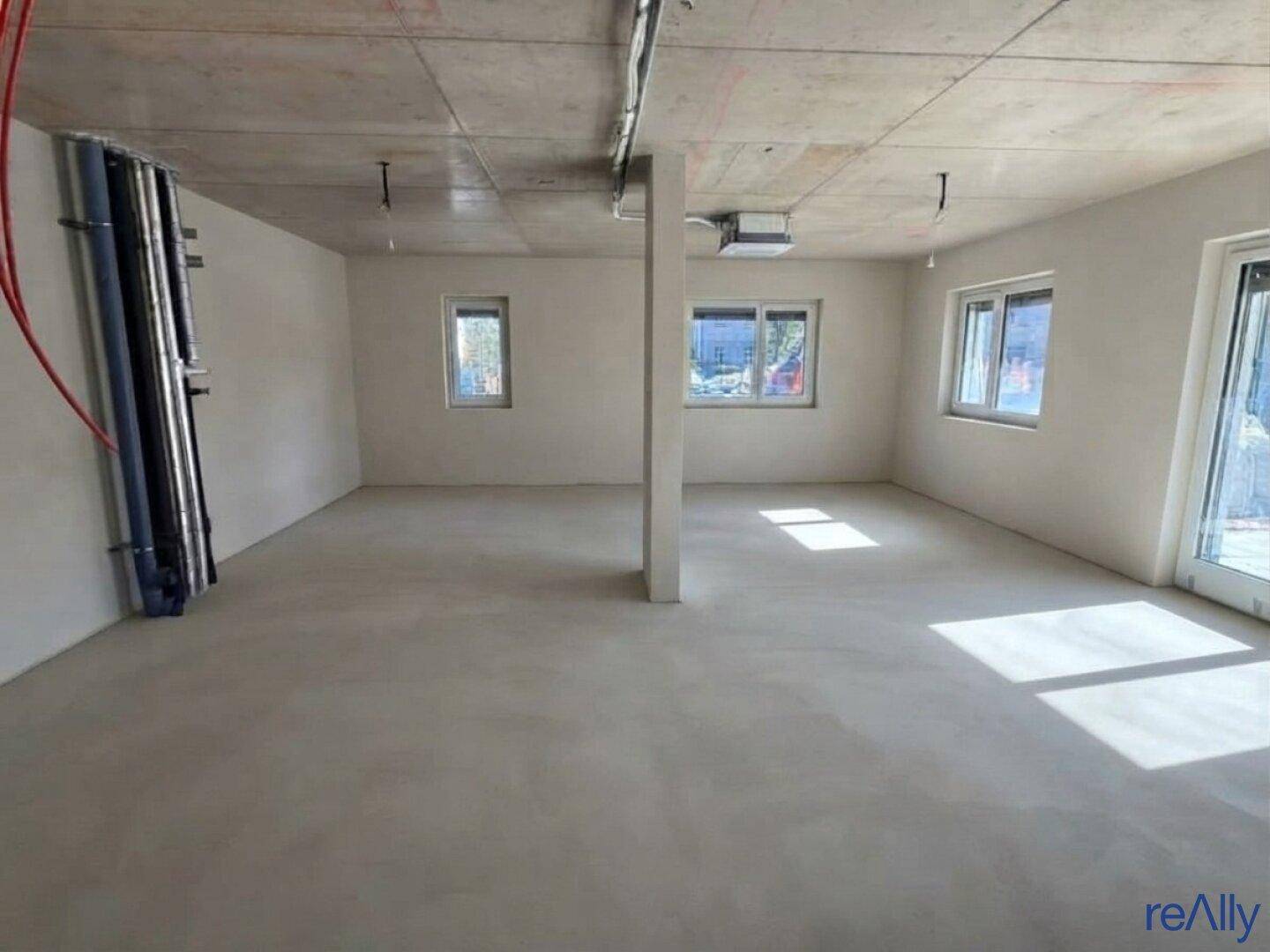 Großer Rohbau mit Betonboden, einer Säule und Fenstern, die Tageslicht hereinlassen.