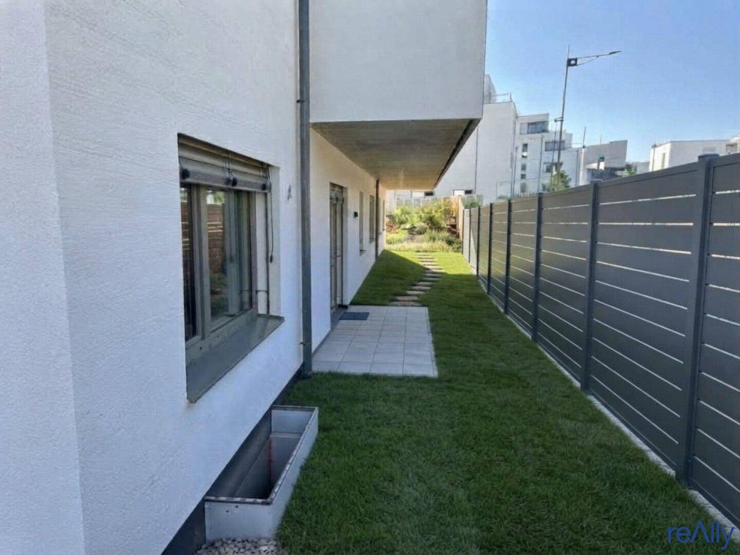 Schmaler Gartenstreifen entlang der Gebäudeseite mit Zaun und einem Fenster mit Rollladen.