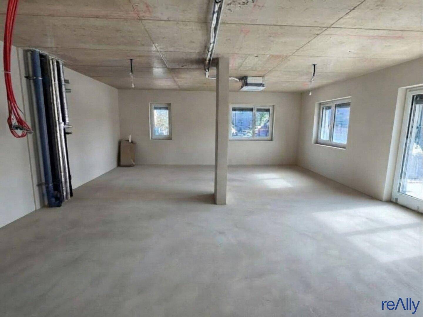 Großer, unfertiger Raum mit Betonboden, sichtbaren Rohren und einem tragenden Pfeiler in der Mitte.
