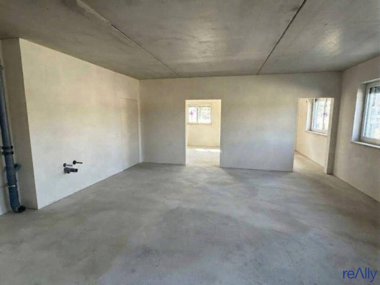 Leerer, unfertiger Raum mit Betonboden und -decke, weißen Wänden und Durchgängen zu weiteren Zimmern.