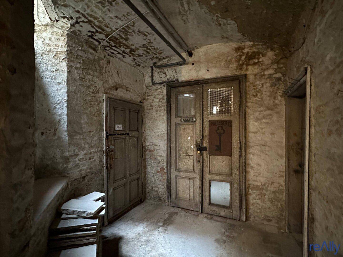 Alter Flur im Keller mit zwei Holztüren und gestapelten Fliesen, der den Charme eines historischen Gebäudes bewahrt.