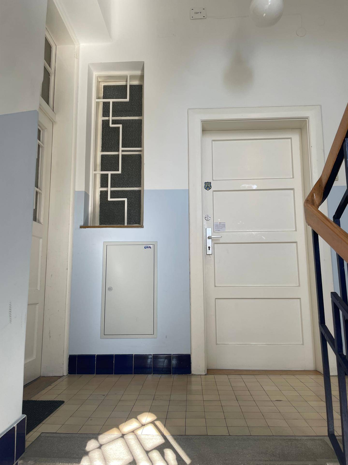 Helles Treppenhaus mit weißer Tür, dekorativem Fenster und Zugang zu den oberen Etagen.