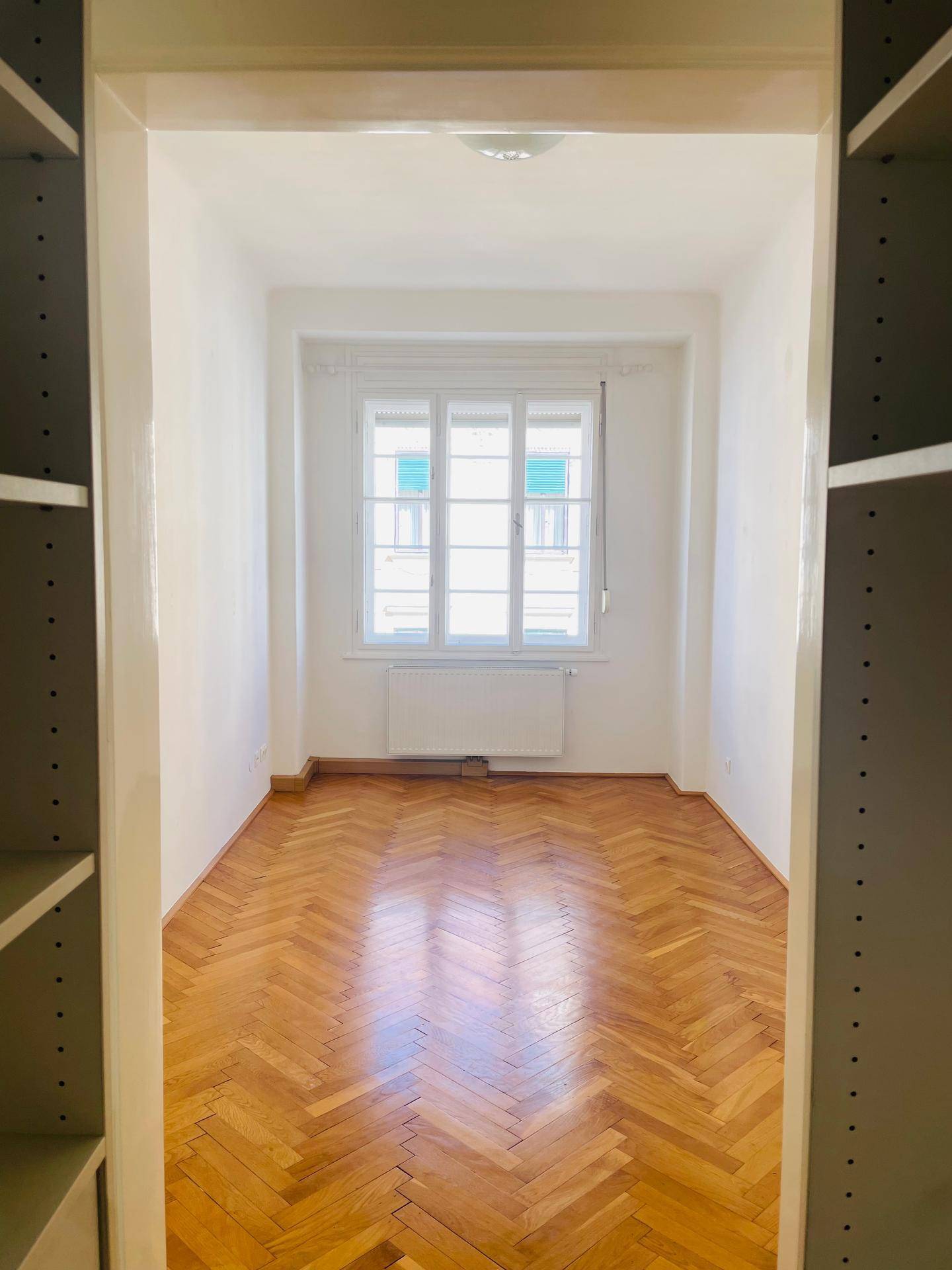 Blick aus einem begehbaren Schrank in ein helles Zimmer mit Fischgrätparkett und Fenster.