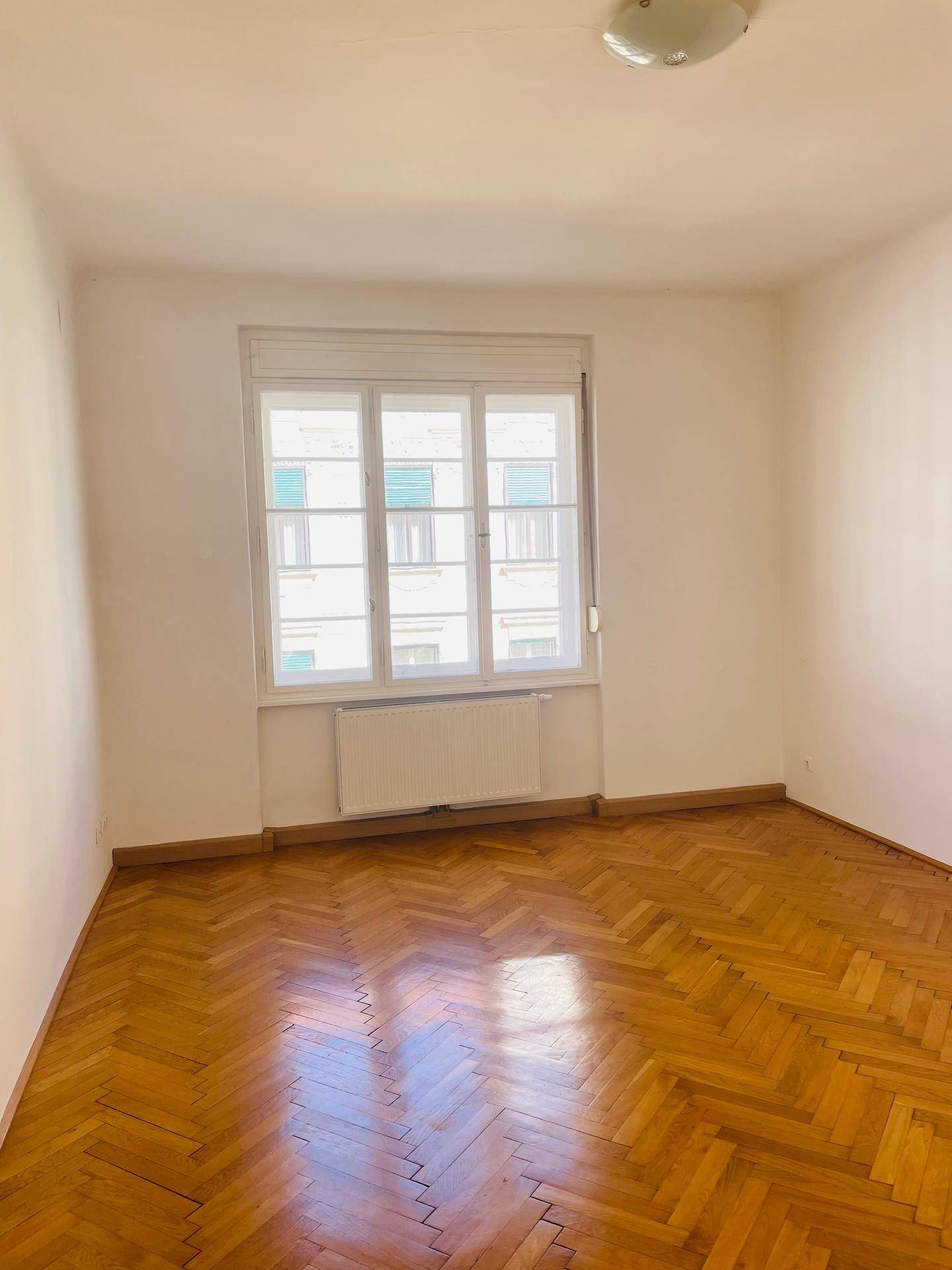 Helles Zimmer mit klassischem Fischgrätparkett und einem Fenster, das für natürliche Belichtung sorgt.