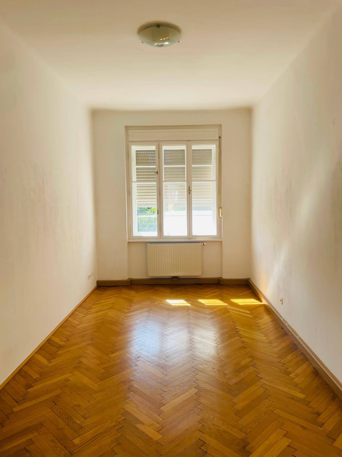 Geräumiges Zimmer mit elegantem Fischgrätparkett und einem großen Fenster mit Jalousien.