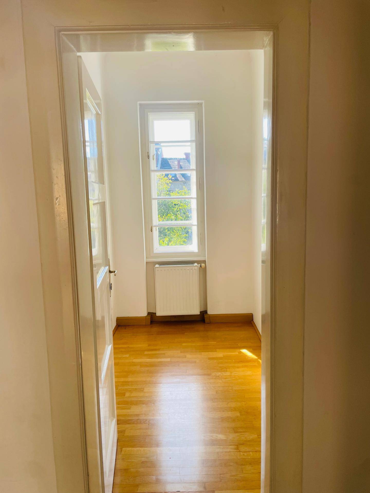 Helles Zimmer mit klassischem Parkettboden und einem hohen Fenster, das viel Licht hereinlässt.