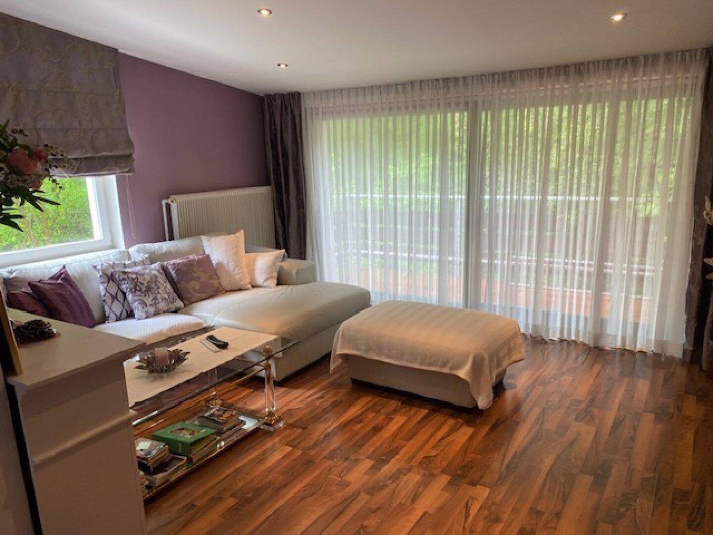 Geräumiges Wohnzimmer mit Holzboden, bequemer Sitzecke und großen Fenstern mit Vorhängen.