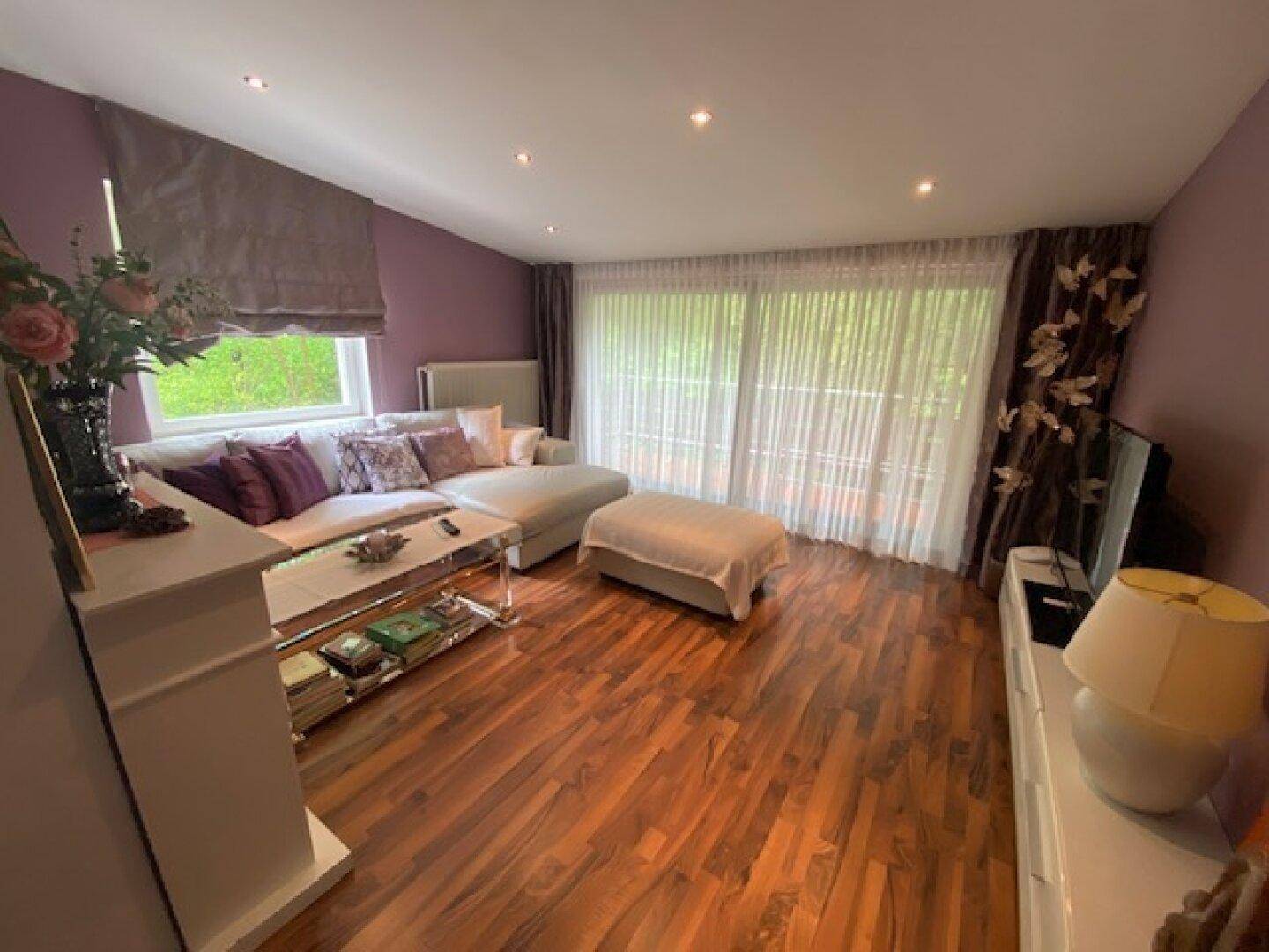 Gemütliches Wohnzimmer mit Holzboden, Ecksofa und großen Fenstern mit Blick ins Grüne.