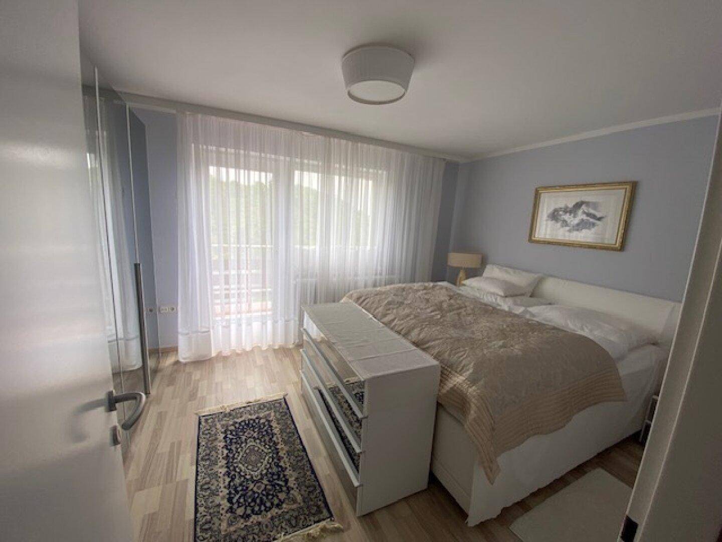Schlafzimmer mit hellem Holzboden, Doppelbett und großen Fenstern mit transparenten Vorhängen.