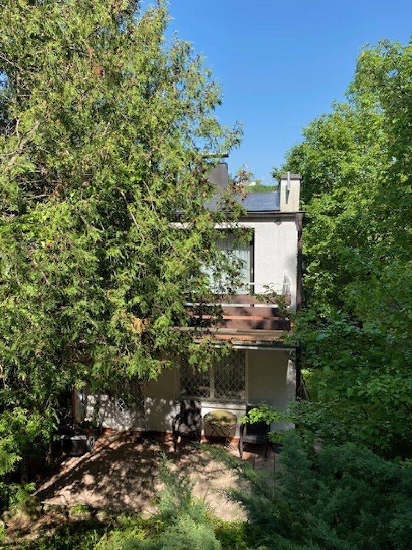 Rückansicht des Hauses mit Balkon und einer kleinen Terrasse im Erdgeschoss, umgeben von Bäumen.
