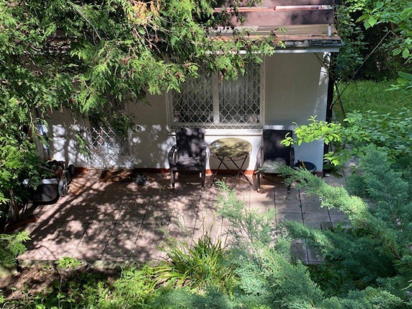 Kleine gepflasterte Terrasse mit Gartenmöbeln, umgeben von Bäumen und Sträuchern.