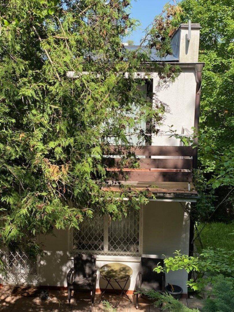 Rückansicht des Hauses mit Balkon und einer kleinen Terrasse im Erdgeschoss, umgeben von Bäumen.