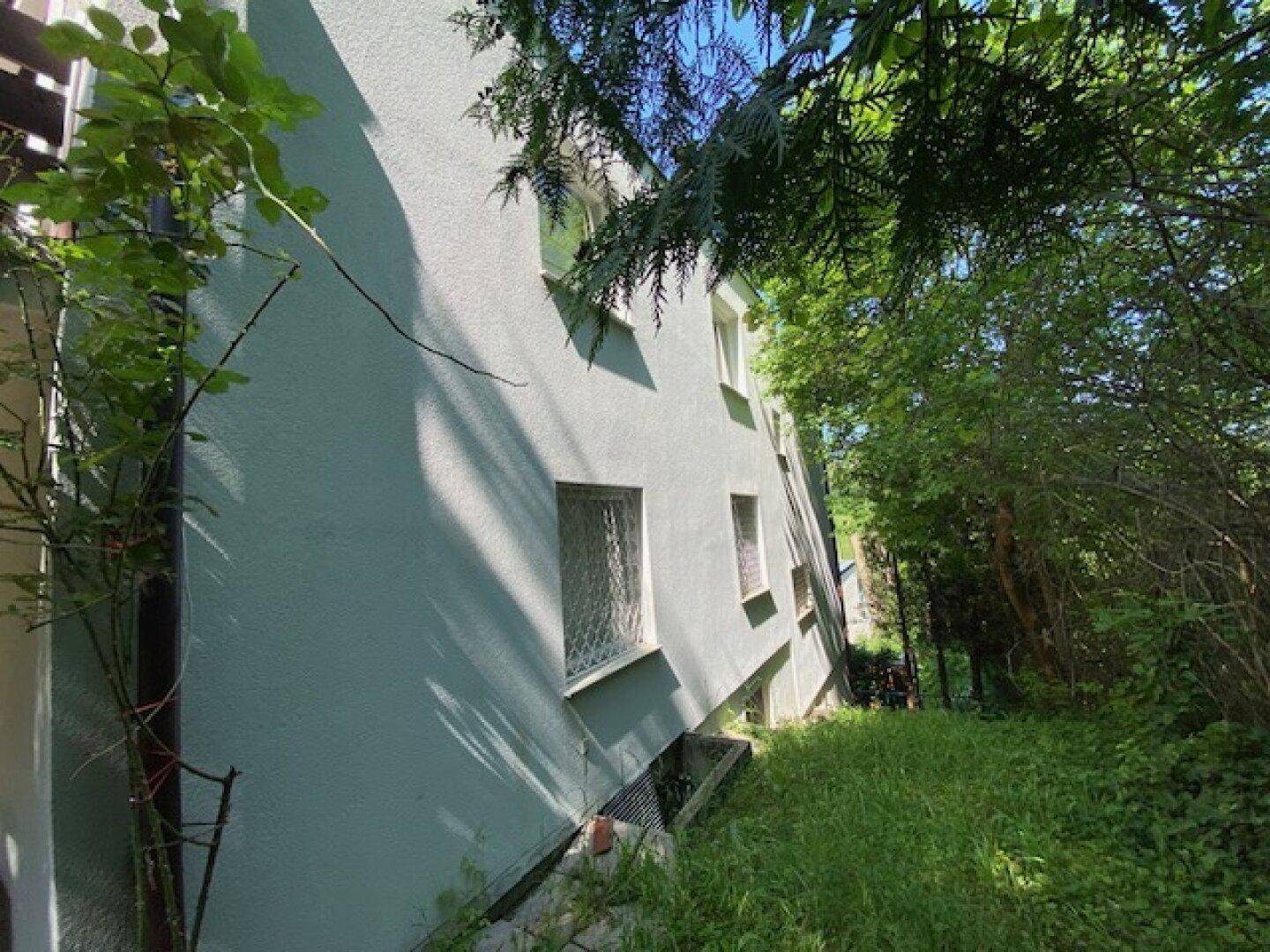 Fassade eines Gebäudes, umgeben von üppigem Grün und Bäumen, mit sichtbaren Fenstern.