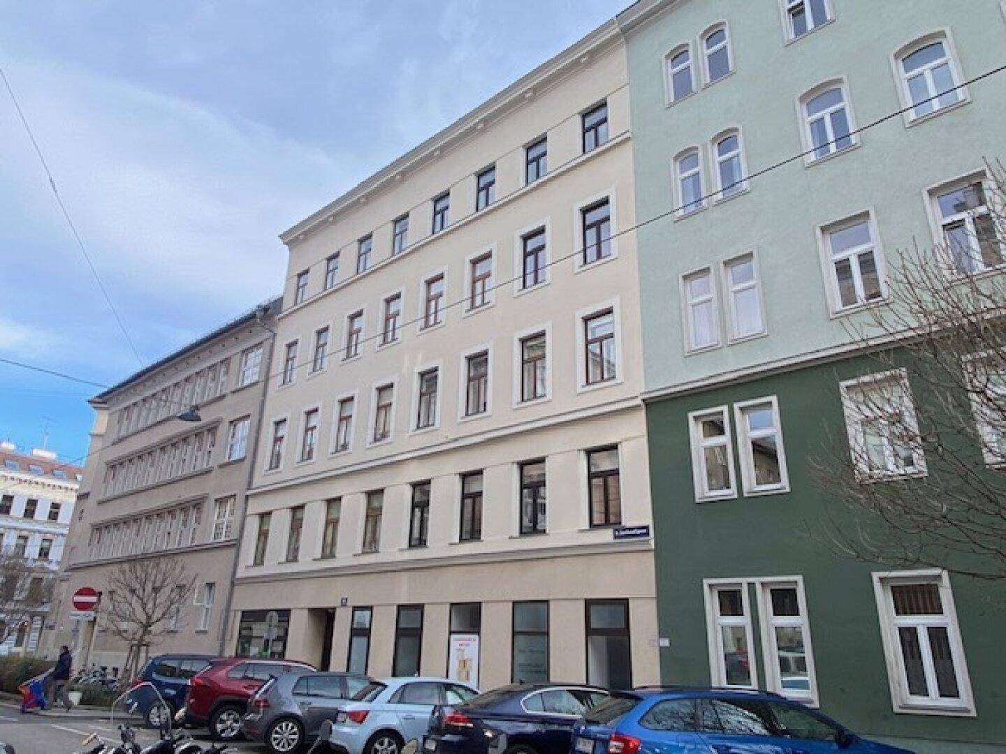 Historische Gebäudefassade an einer Stadtstraße mit vielen Fenstern und parkenden Autos davor.