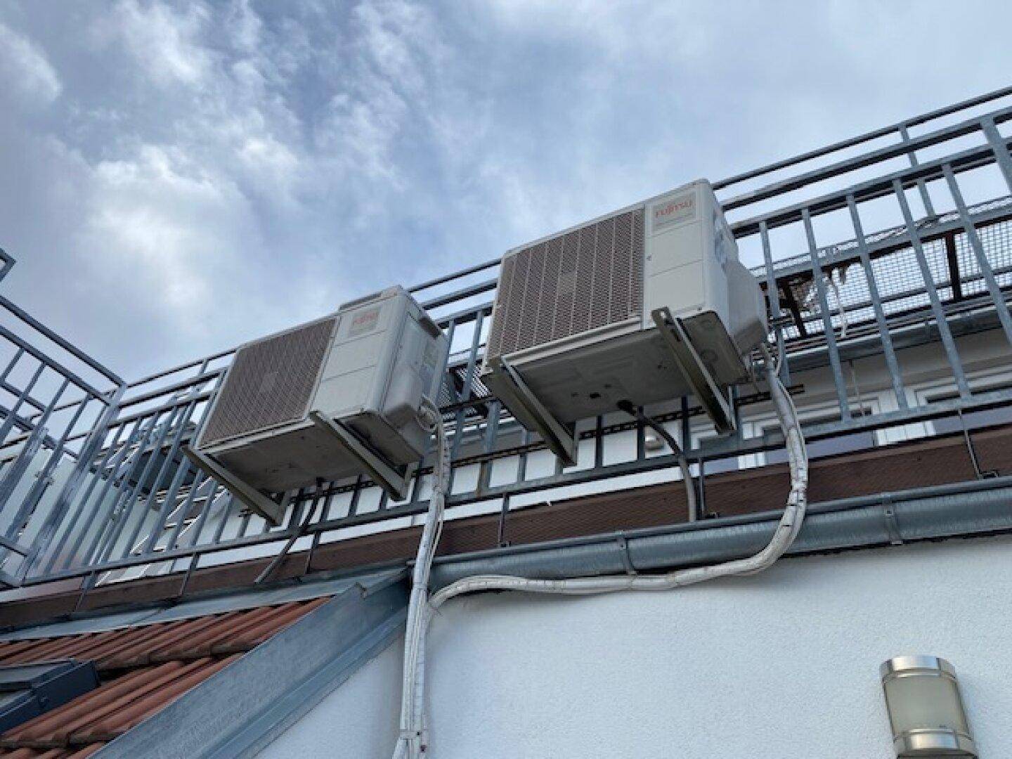 Zwei Klimaanlagen-Außengeräte an der Fassade mit Metallgeländer und Ziegeldach im Hintergrund.
