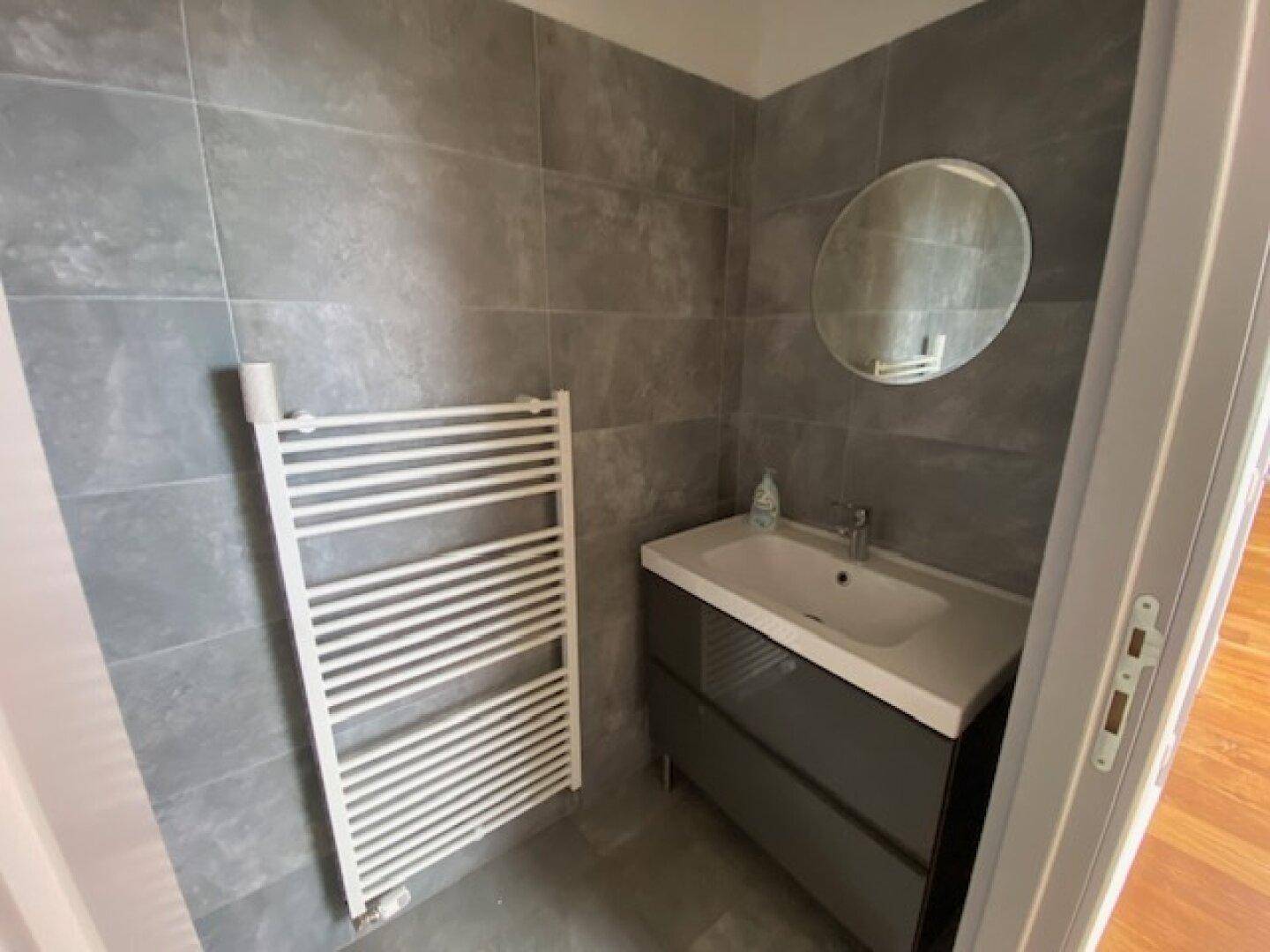 Kleines Badezimmer mit Waschtisch, rundem Spiegel und Handtuchheizkörper, alles in modernen Grautönen.