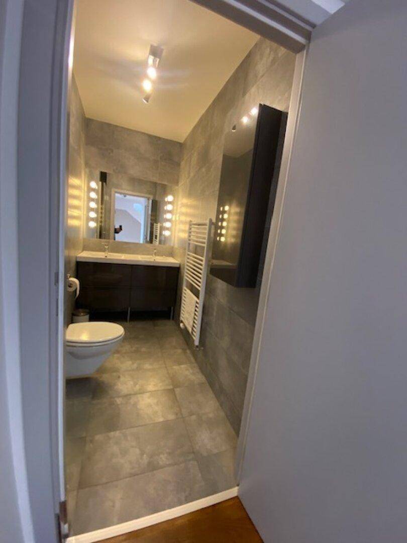 Modernes Badezimmer mit grauen Fliesen, wandhängendem WC, Waschtisch und beleuchtetem Spiegel.