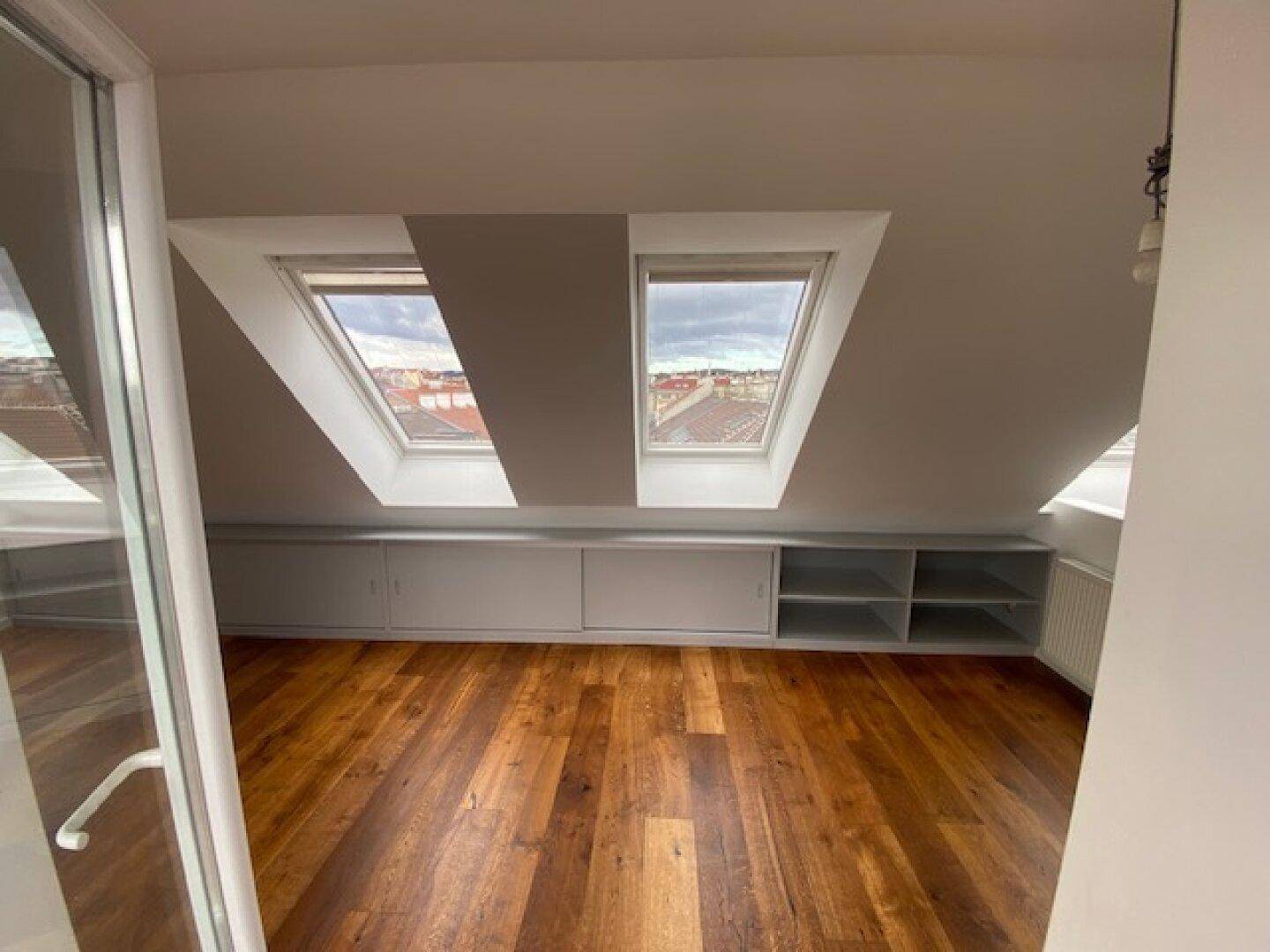 Dachgeschossraum mit Holzboden, zwei Dachfenstern und integrierten Schränken unter der Schräge.