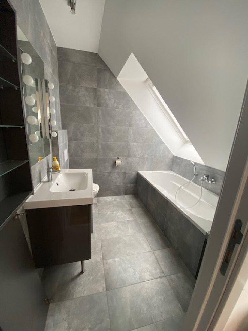 Helles Badezimmer mit Badewanne, Waschtisch und modernen grauen Fliesen im Dachgeschoss.