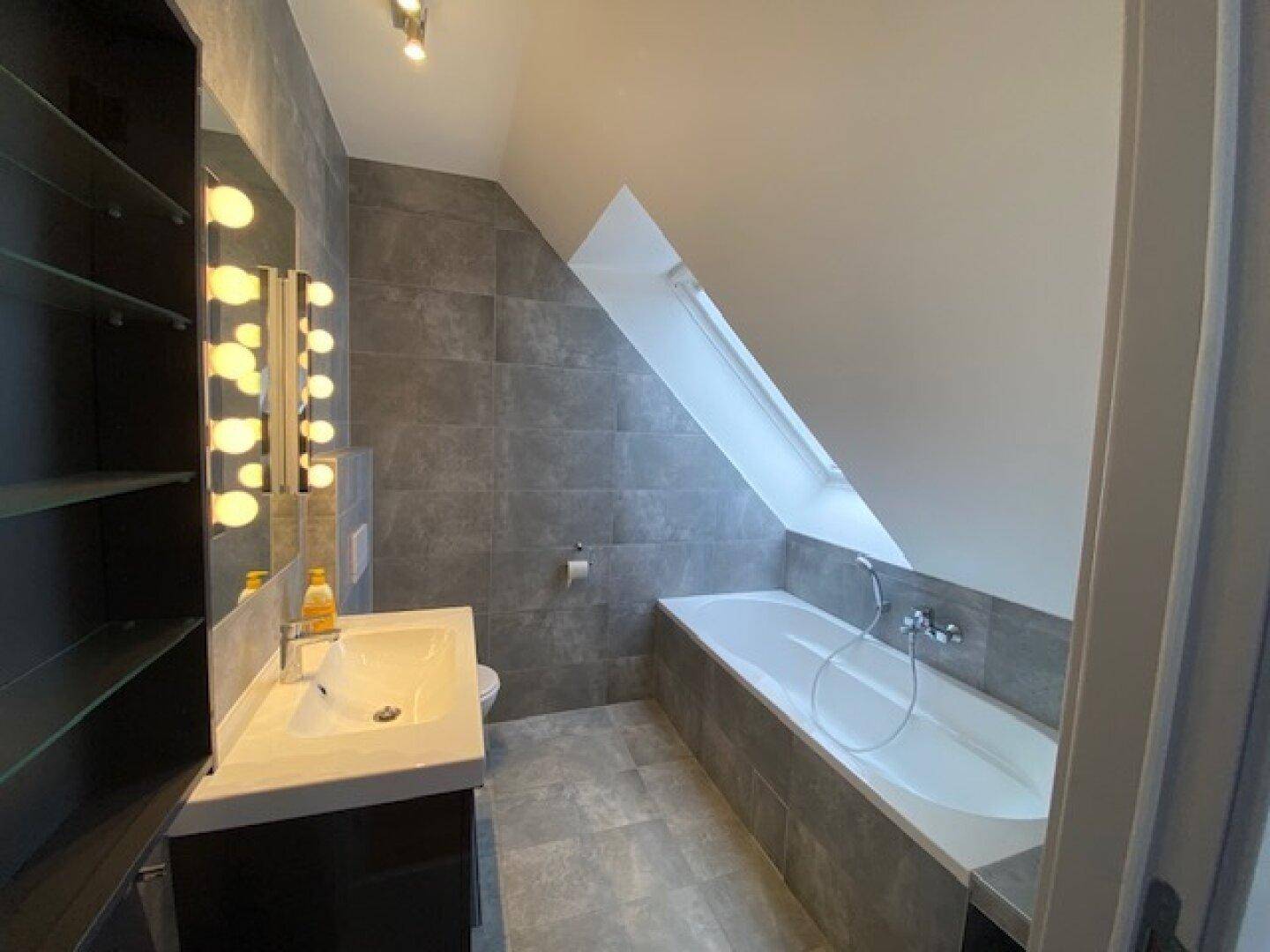Modernes Badezimmer mit grauen Fliesen, Badewanne unter der Dachschräge und Waschtisch.