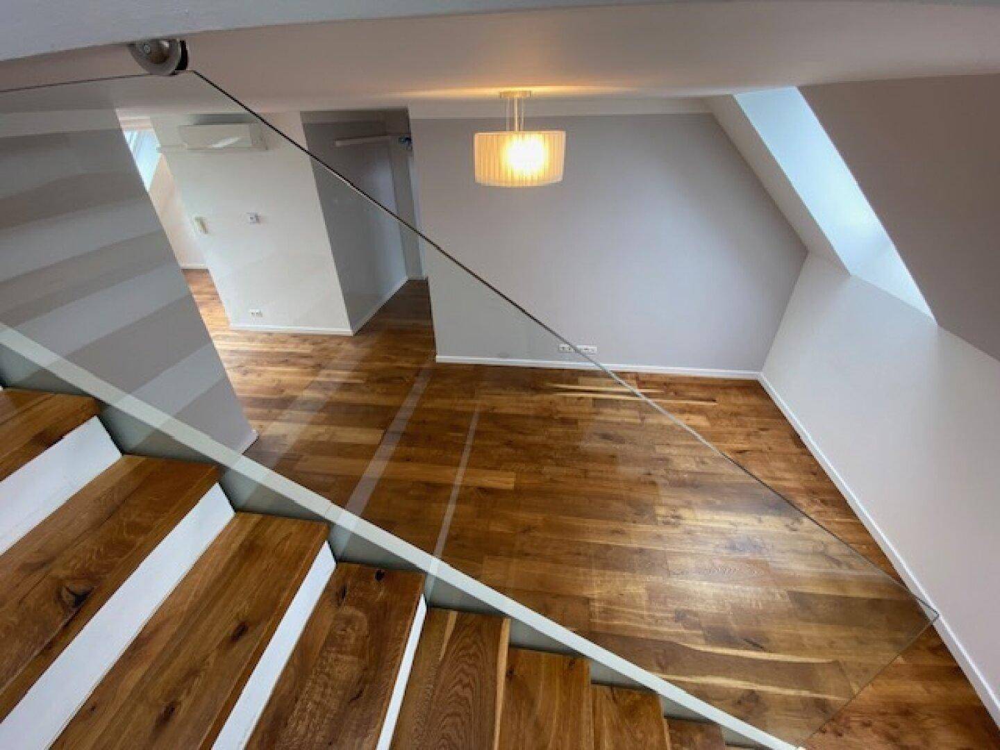Heller Innenraum mit Holztreppe, Glasgeländer und hochwertigem Parkettboden unter Dachschrägen.