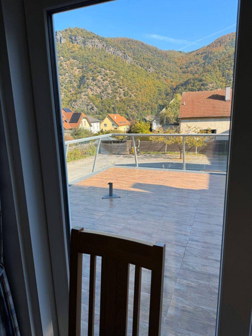 Blick aus einem Fenster auf eine Außenterrasse mit Bergblick unter blauem Himmel.