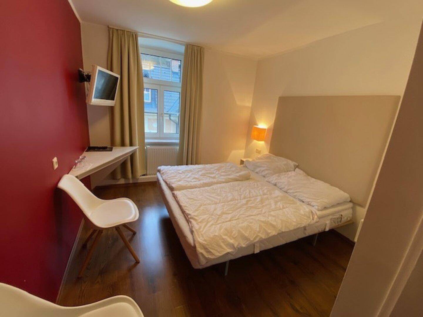 Gemütliches Schlafzimmer mit Doppelbett, Holzboden und roter Akzentwand, hell durch Fenster.