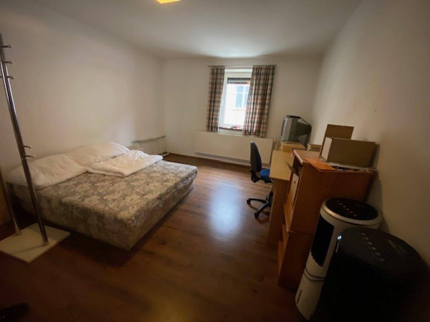 Einfaches Schlafzimmer mit Bett, Schreibtisch, Stuhl und Fenster mit karierten Vorhängen.