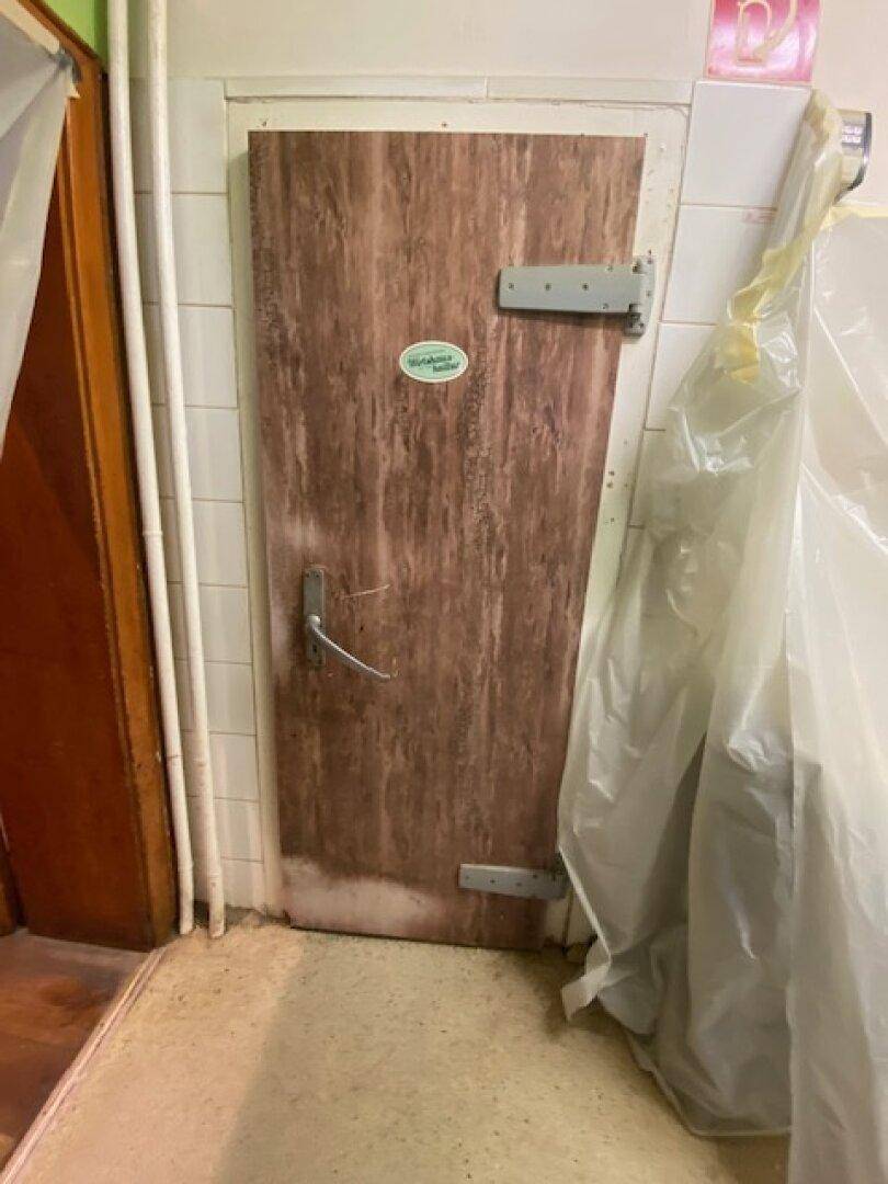Eine Holztür mit Metallbeschlägen in einer gefliesten Wand, möglicherweise zu einem Kühlraum führend.