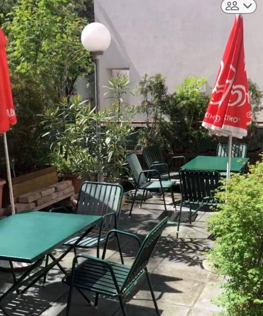 Grüne Außenterrasse mit Tischen und Stühlen unter Sonnenschirmen, umgeben von Pflanzen.