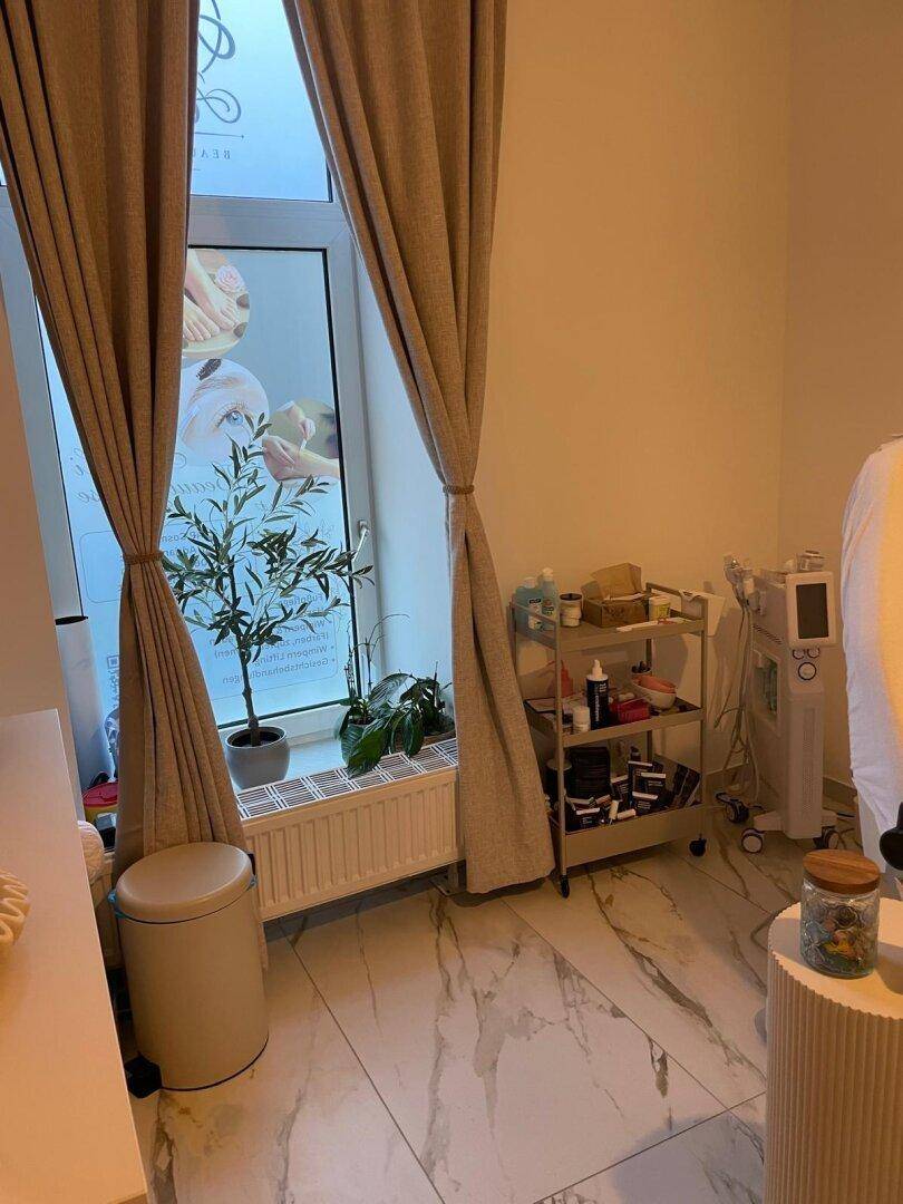 Fensterbereich eines Behandlungsraums mit beigen Vorhängen, Heizkörper und einer kleinen Pflanze.