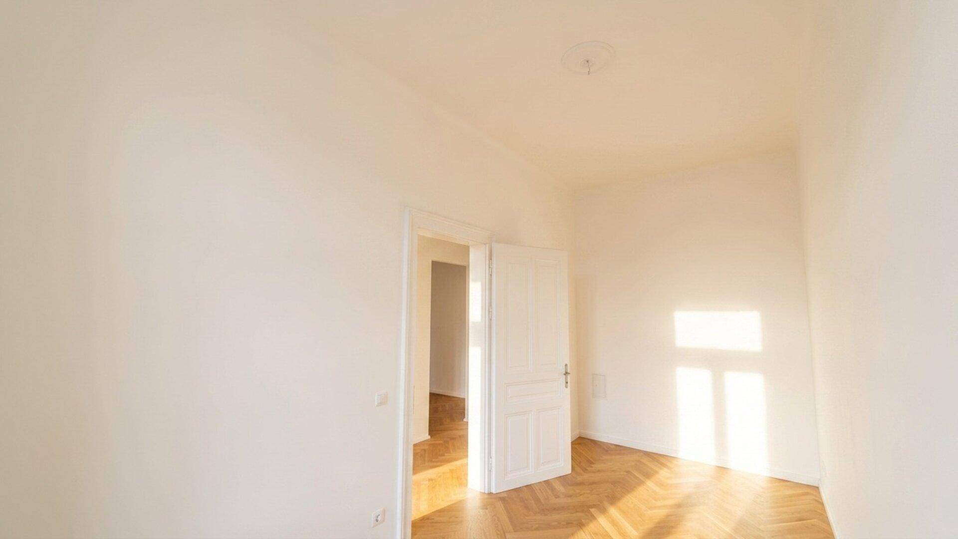 Heller Raum mit Fischgrätparkettboden und weißen Wänden, durch dessen Fenster Sonnenlicht einfällt.