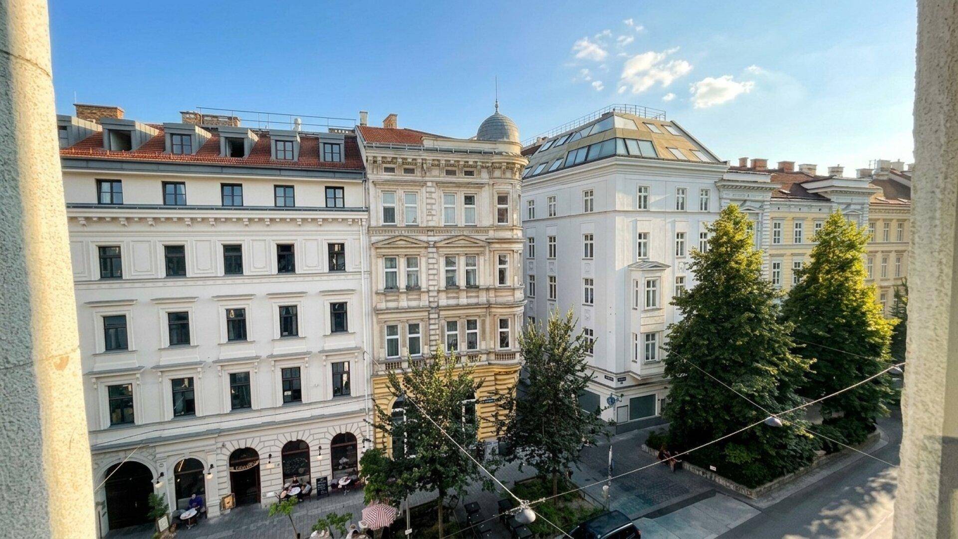 Blick aus dem Fenster auf eine belebte Straße mit historischen Gebäuden und grünen Bäumen unter blauem Himmel.