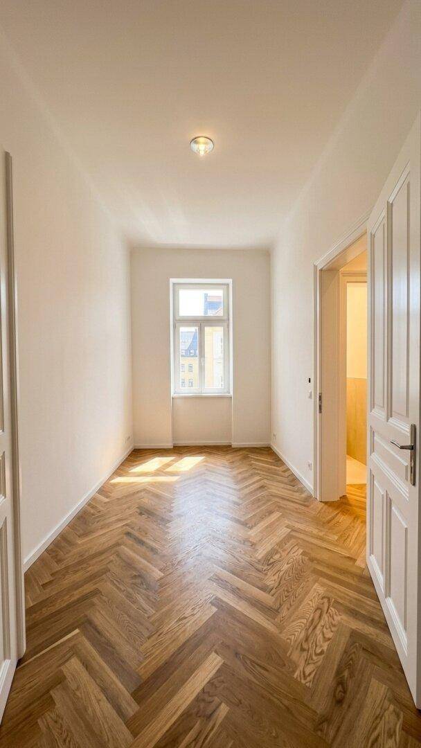 Heller Raum mit Fischgrätparkettboden, weißen Wänden und einem Fenster am Ende des Zimmers.