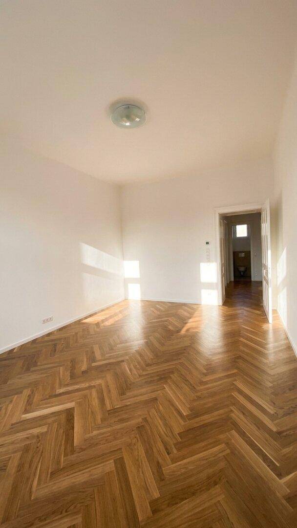 Geräumiger, heller Raum mit klassischem Fischgrätparkett und viel Sonnenlicht, das durch die Fenster fällt.