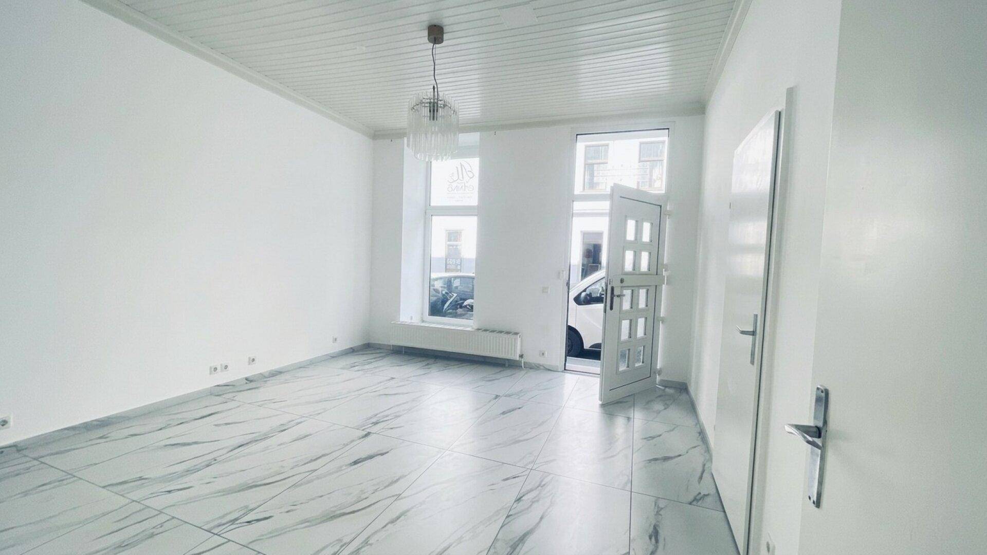 Heller, leerer Raum mit modernen Marmorfliesen, hohen Decken und einer geöffneten Eingangstür zur Straße.