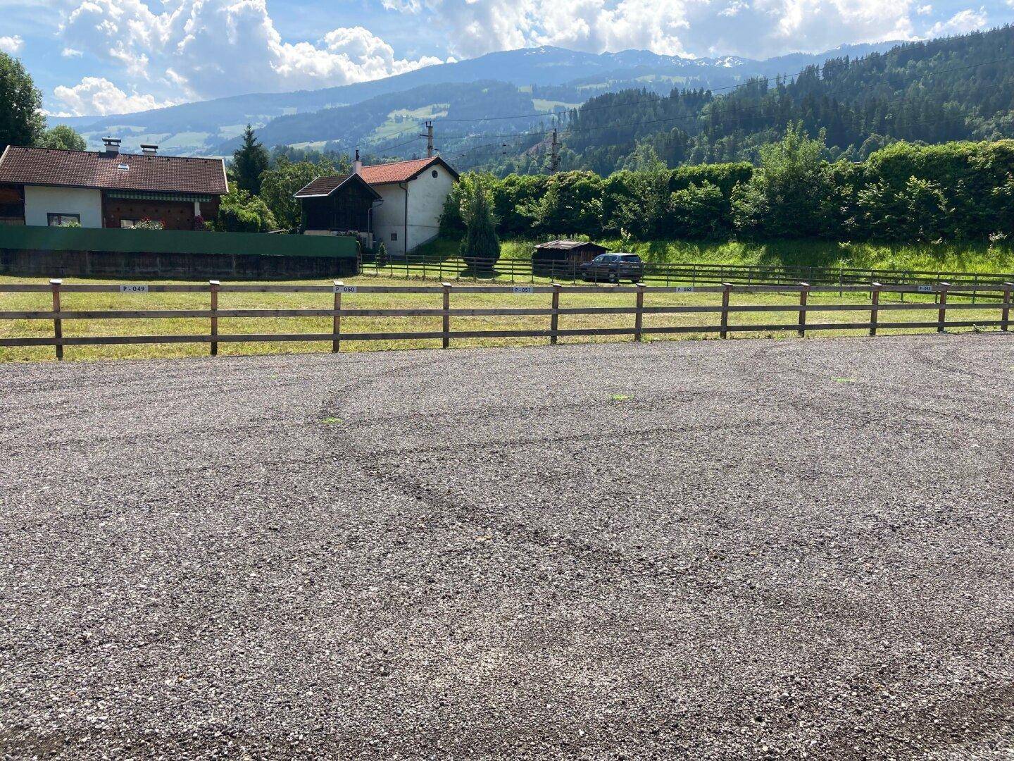 Weitläufiger Schotterbereich mit Holzzaun, umgeben von grüner Natur und Bergen im Hintergrund.