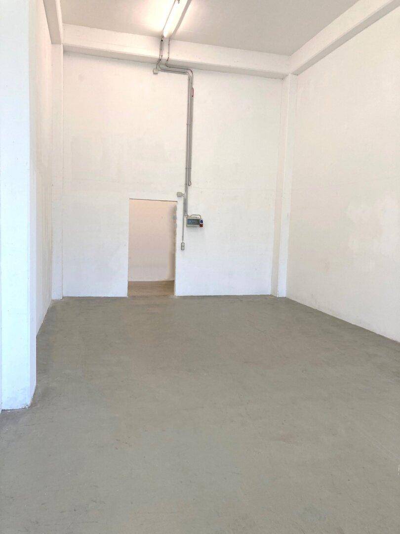 Geräumiger, leerer Gewerberaum mit Betonboden, hohen Decken und sichtbaren Leitungen an der Wand.