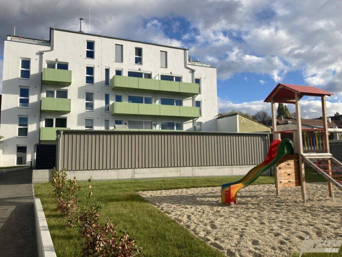 Rückansicht des Neubaus mit Spielplatz und gepflegter Grünfläche unter blauem Himmel.