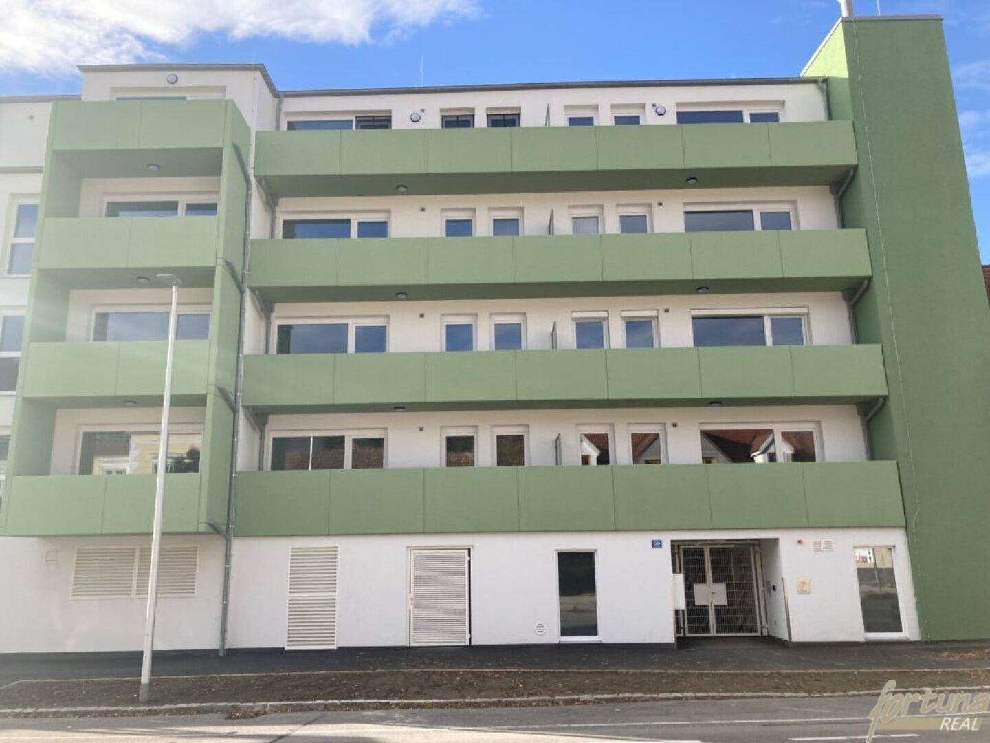 Modernes Mehrfamilienhaus mit grünen Balkonen und weißer Fassade unter blauem Himmel.