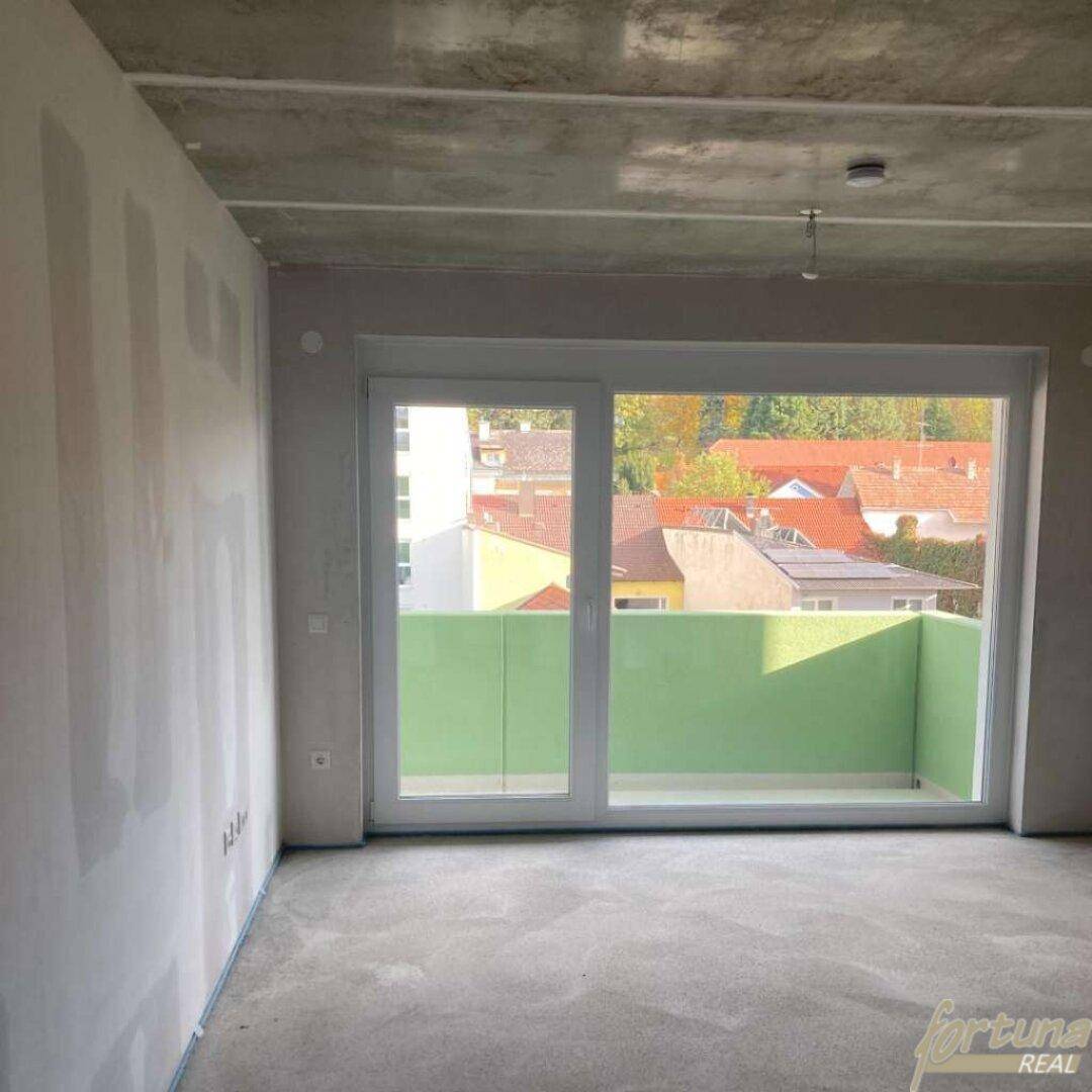 Unfertiger Innenraum mit großem Fenster und Zugang zu einem Balkon, noch im Bauzustand.